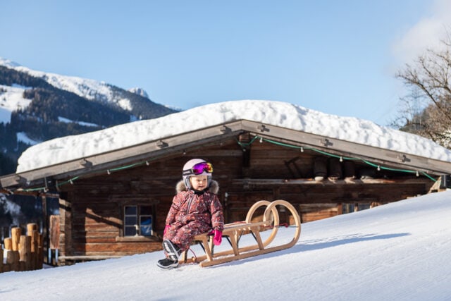 Kind mit Schlitten im Schnee vor Hütte.